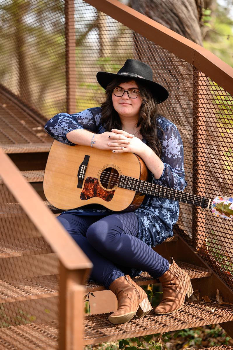 Charity '24 standing on the Brushy Fork copper bridge step. She is holding a guitar on her lap.