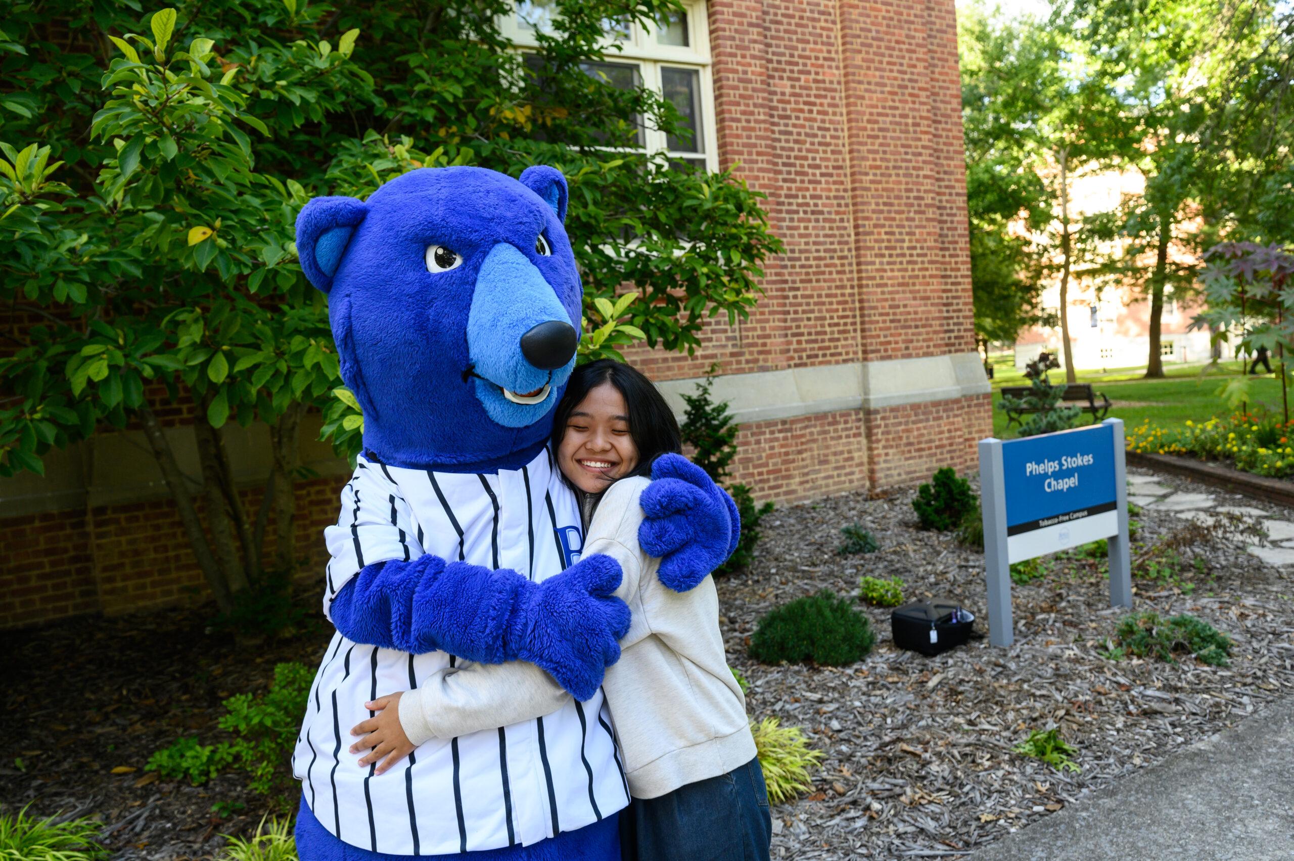 Photo of a student hugging Berea's mascot, Blue, a blue bear outside. A green tree and brick building are in the background.