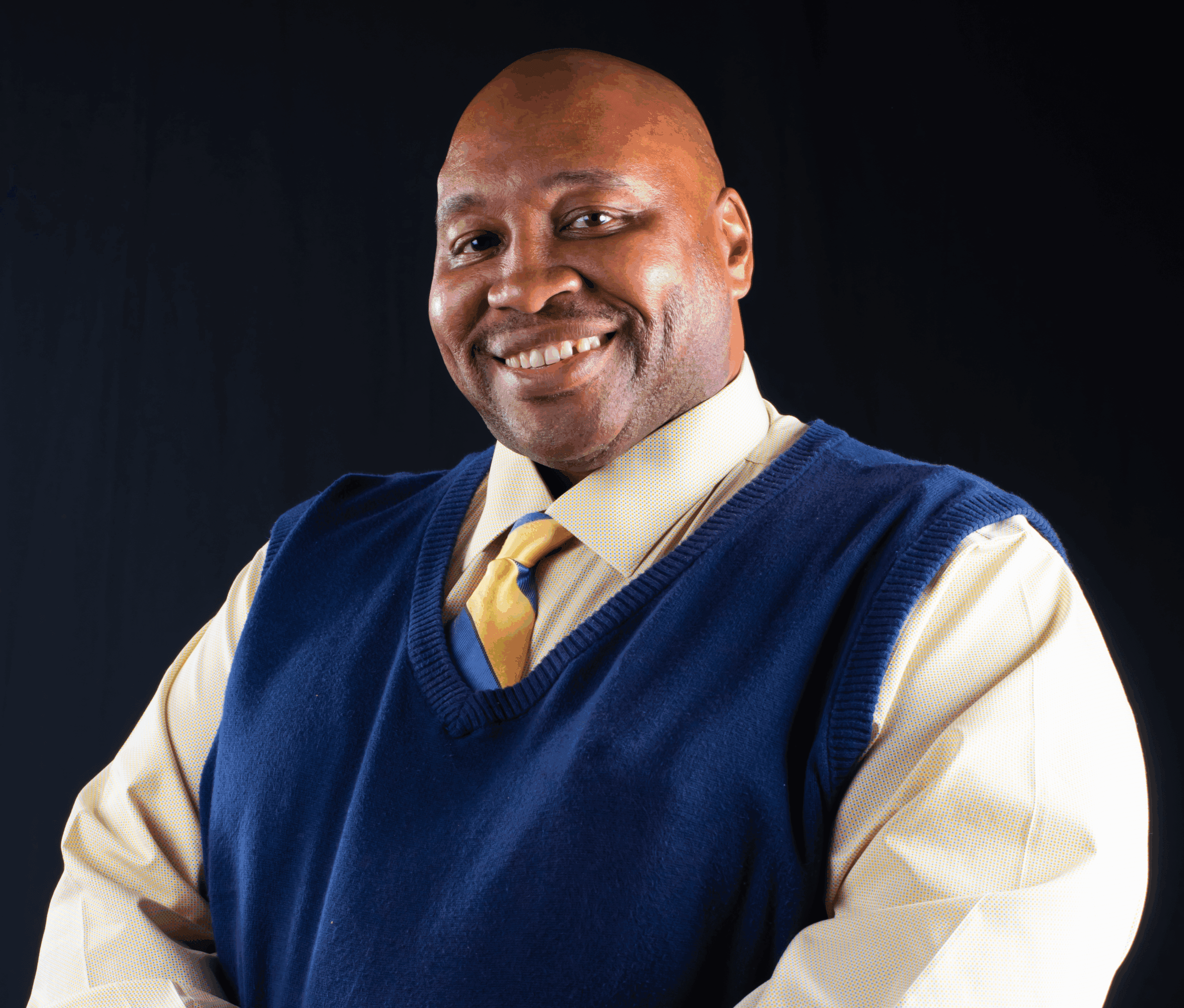 A photo of Senior Regional Admissions Manager for the Tri-City region, Frank Polion, standing against a black background, wearing a yellow long-sleeved yellow shirt, a blue an yellow striped tie, and a navy blue swear, smiling.